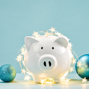 A white piggy bank sitting in front of blue background covered with holiday lights and ornaments. Overcome Holiday Impulse Purchases