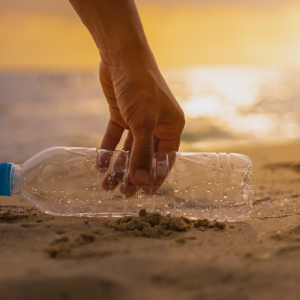 A hand picking up an empty plastic bottle on the beach at sunset