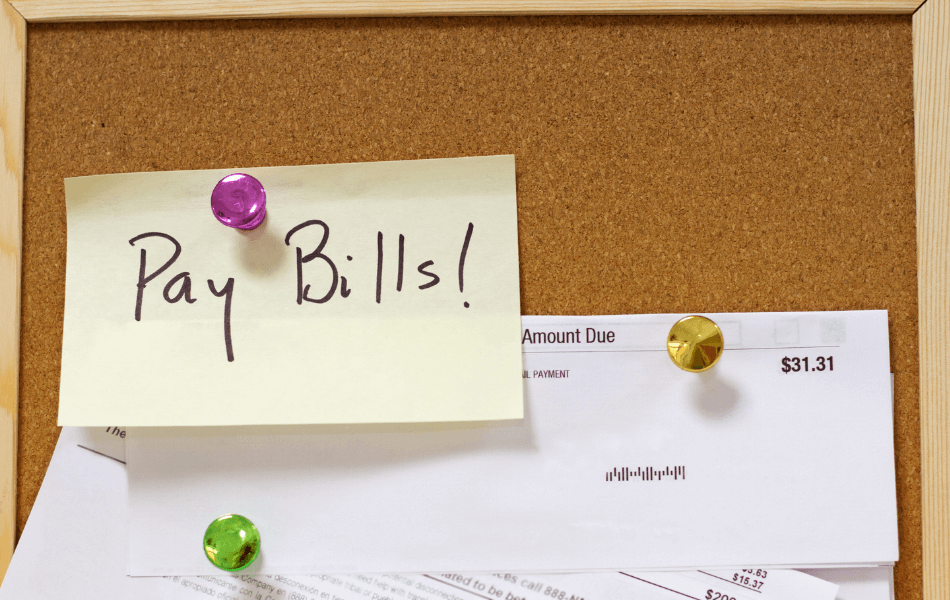 Sticky note that says "Pay Bills" attached to a bulletin board.